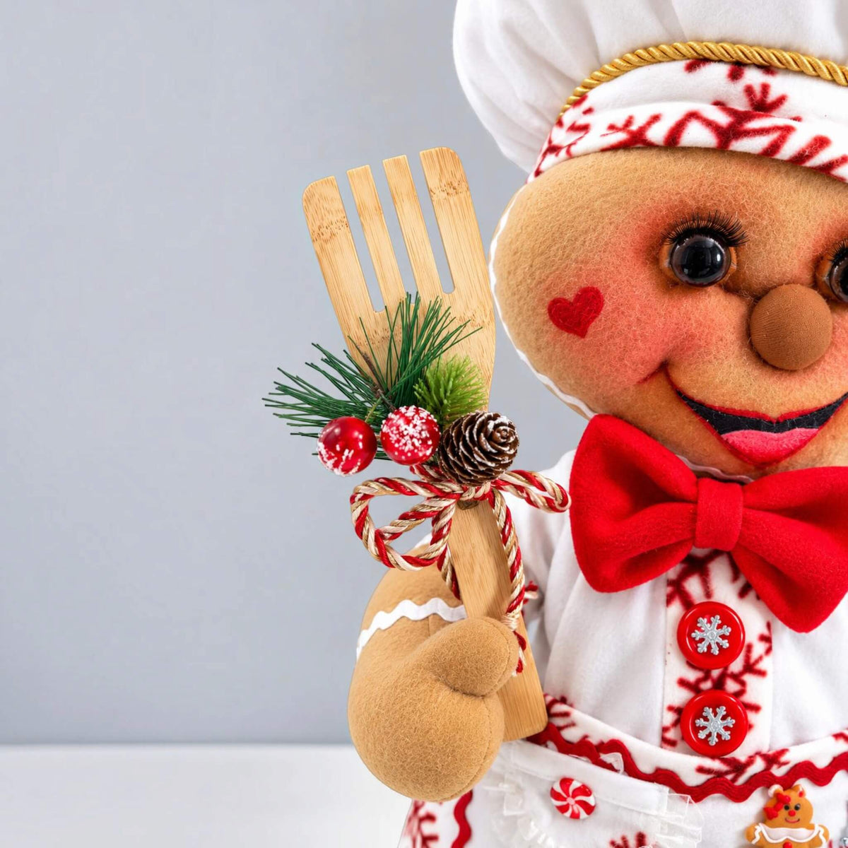 Close-up of handmade Christmas Gingerbread Baker holding a wooden baking utensil decorated with greenery, berries, pinecone, and ribbon.