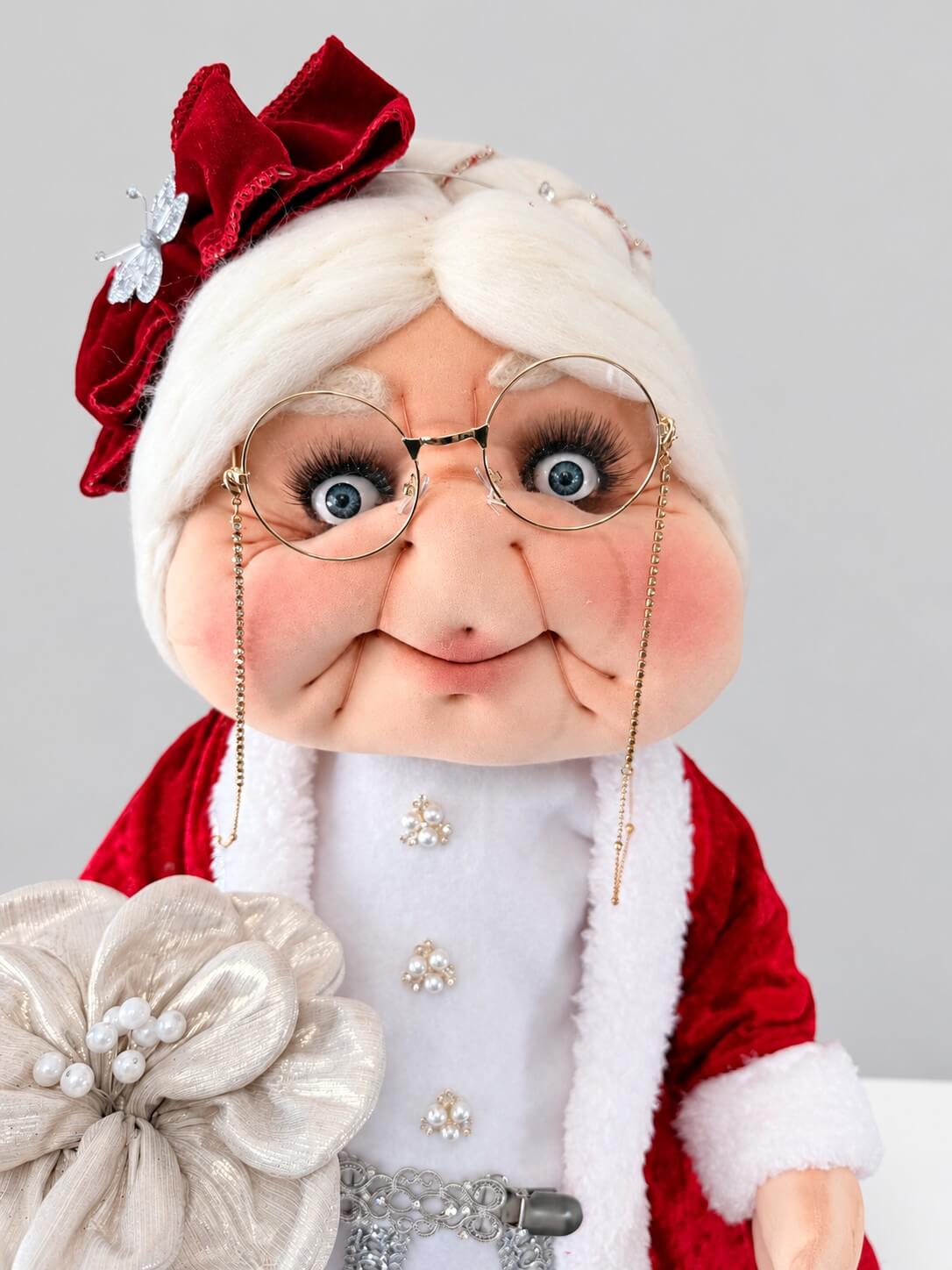 Close-up of handmade Mrs. Claus with white hair, glasses, rosy cheeks, a red bow, and a silver flower.
