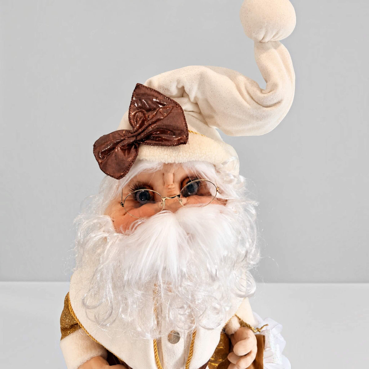 Close-up of gold Santa Claus’s face, glasses, white beard, and cream hat with a brown bow.